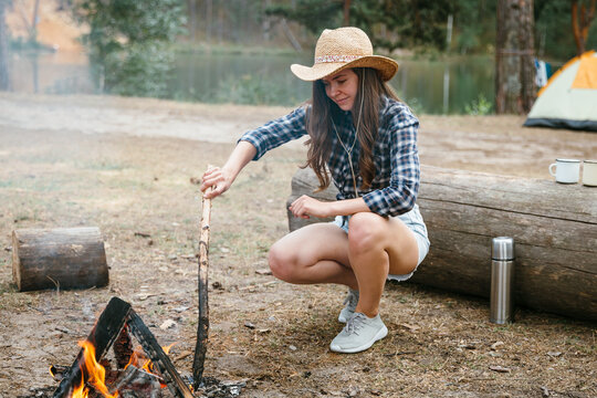 Woman in a straw hat tends to a campfire at a forest campsite near a lake, outdoor adventure story, relaxing nature getaway, peaceful camping lifestyle.