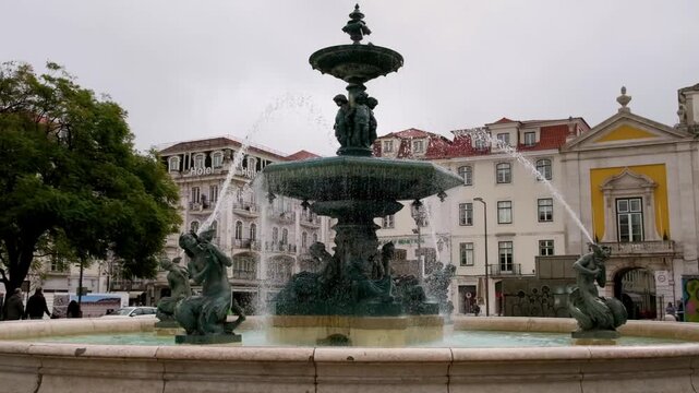 The ornate Rossio Square fountain stands in central Lisbon, Portugal, surrounded by historic buildings and patterned pavement under a cloudy sky