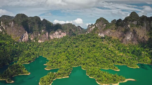 Explore beauty of Cheow Lan Lake in Khao Sok National Park, Thailand. Lush tropical rainforest, towering limestone karst mountains and vibrant emerald water under bright sky. Aerial view drone shot