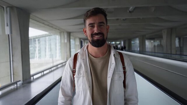 Handsome young man with a backpack smiling at the camera while standing on a moving walkway in an airport terminal.