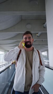 Man at airport moving walkway using phone, talking on a cheerful business conversation. Vertical video with copy space.