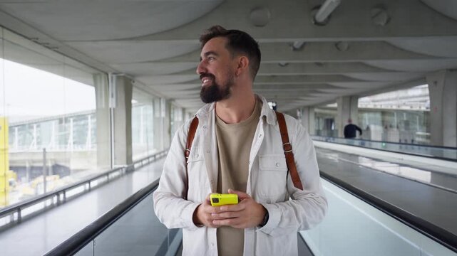 Smiling traveler with a backpack texting on his phone while riding an airport travelator