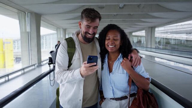 Happy multiracial couple with backpacks using phone on a moving walkway in an airport terminal going on vacation.
