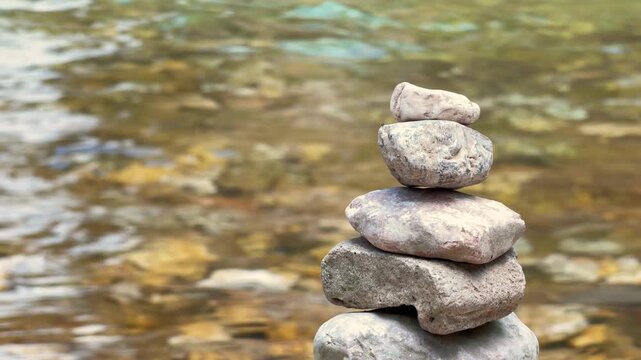 A stack of balanced stones stands on a riverbank with clear shallow water rippling behind it, a calm concept of zen balance and mindfulness.