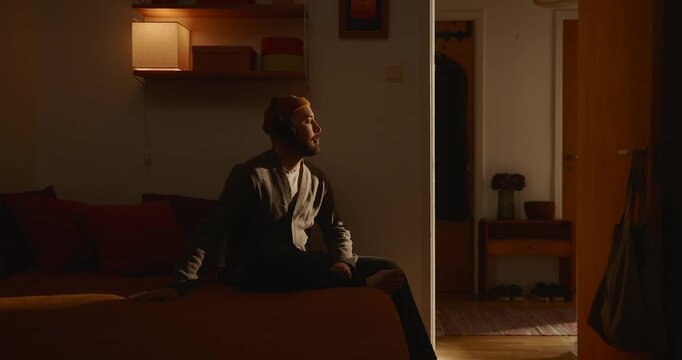 Young man wearing headphones sitting on bed with eyes closed enjoying music in cosy warm ambient lighting evening room peaceful home interior relaxed mood atmosphere hygge quiet leisure lifestyle