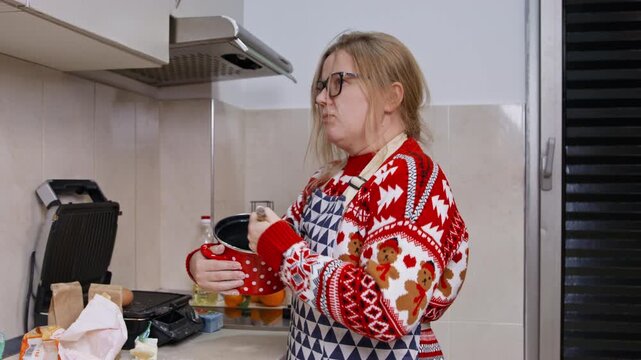 An adult woman wearing glasses and a festive red sweater with an apron stirs a pot on a kitchen counter, preparing a holiday meal at home.