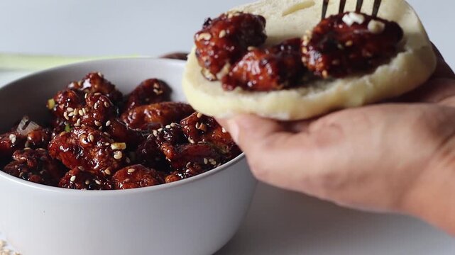 Close up video of honey glazed sesame chicken being placed into ciabatta bread using a fork, showing food preparation and sandwich assembly in a styled kitchen setting