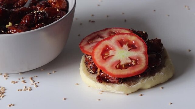 Close up video showing fresh tomato slices being added to honey glazed sesame chicken on ciabatta bread, followed by placing the top half to complete the sandwich