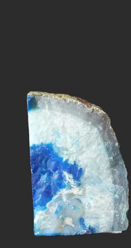 Polished blue agate slice rotating against a dark gray background, showing vivid blue and white concentric bands, translucent mineral layers and a rough natural outer edge.