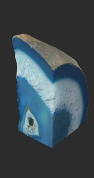 Vertical view of a blue and white banded agate section rotating on a dark gray background, showing concentric mineral patterns, polished surfaces and a contrasting rough exterior.