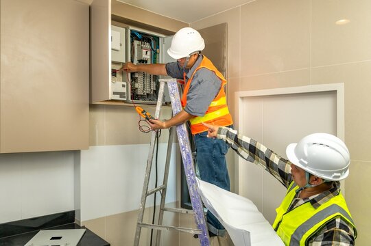 Detailed view of two electricians working on electrical panel installation with safety gear and tools Electricians using clamp meter inspecting electrical panel board with blueprint on ladder