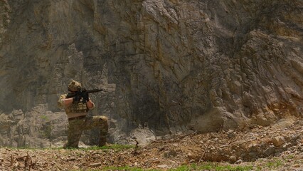 Fototapeta premium A special forces soldier in combat gear kneels with a rifle at the base of a cliff, maintaining vigilance during a tactical operation in a rugged environment.