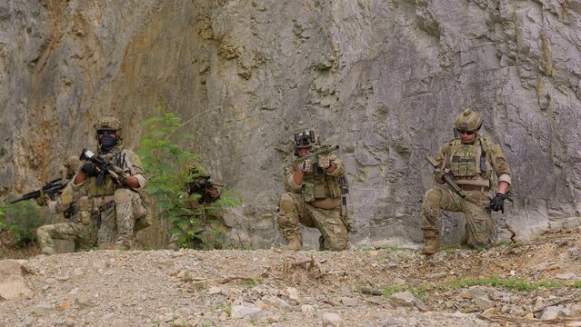 A special forces team conducts a military operation, with soldiers aiming rifles from defensive positions against a rugged rock face background.