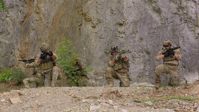 A special forces team conducts a military operation, with soldiers aiming rifles from defensive positions against a rugged rock face background.