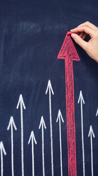 Hand drawing red arrow upwards on chalkboard, representing progress, growth, and success among white arrows, symbolizing leadership or achievement.