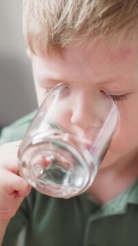 Close up of young boy drinking water from clear glass. child holds transparent mug bringing it to his mouth and taking sips against soft indoor lighting. Background remains blurred