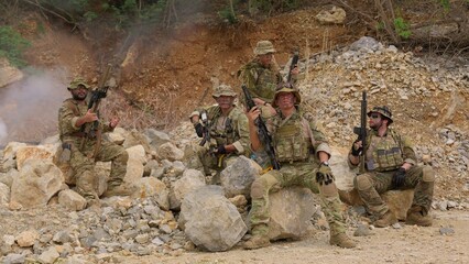 Fototapeta premium An elite team of special forces soldiers in full tactical gear pose together with their rifles amidst smoke and rocks during a military operation in a rugged combat zone.