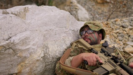 Fototapeta premium An exhausted soldier with face paint rests behind a rock, holding his rifle and catching his breath during a tactical military mission in a rugged environment.