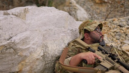 Fototapeta premium An exhausted soldier with face paint rests behind a rock, holding his rifle and catching his breath during a tactical military mission in a rugged environment.