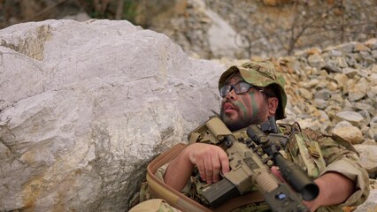 Naklejka premium An exhausted soldier with face paint rests behind a rock, holding his rifle and catching his breath during a tactical military mission in a rugged environment.