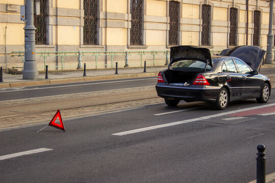 Black sedan with open trunk and hood parked on a city street beside a warning triangle, apparent breakdown scene.