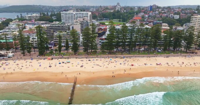 Manly Beach in Sydney, Australia, showing the popular coastline with many people enjoying the sand, swimming in the ocean waves, and the urban cityscape backing the shore