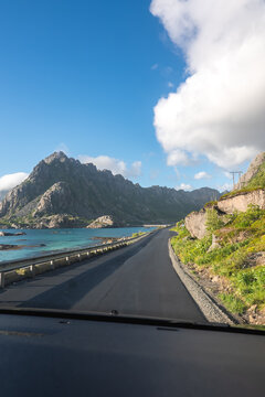 A vehicle drives along an asphalt road next to turquoise water and rocky mountains under a summer sky in the Lofoten islands of northern Norway.