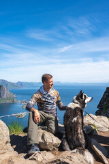 A hiker sits on rocky summit of Reinebringen beside his husky dog, looking at Reine village and Norwegian fjord waters in Lofoten during sunny summer. © Aerial Film Studio