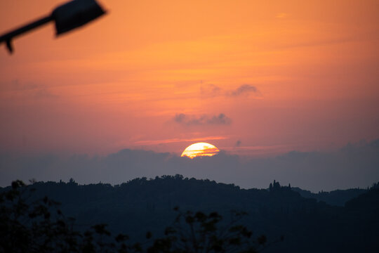 A captivating sunset over rolling hills and mountains in Greece, with the sun partially obscured by clouds, casting a warm orange glow.