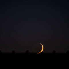 A stunning golden sliver of a crescent moon glows warmly just above the horizon, emerging from the deep darkness of the night sky. An atmospheric and mysterious celestial scene © AgioiDouloi