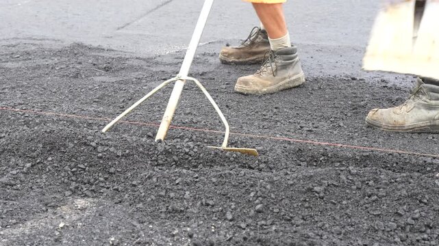 Construction worker using a rake for manual leveling and fine grading of asphalt subgrade layer alongside a reference guide string