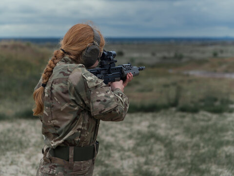 Close view of a red haired servicewoman in camouflage aiming an SA80 rifle downrange, ear defenders and safety glasses correctly worn.