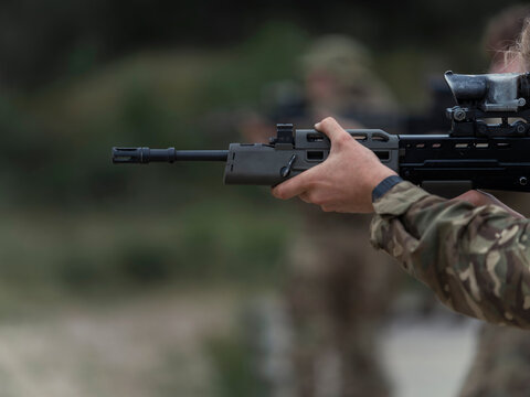 Close side view of a soldier's hand gripping the handguard of a British service rifle, a second shooter blurred in the background.