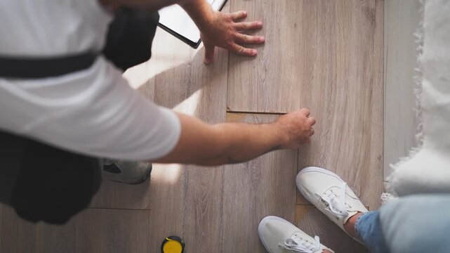 Handyman discussing damaged laminate flooring with female homeowner inside her house, examining the broken floor and discussing repair options, costs and maintenance needs.