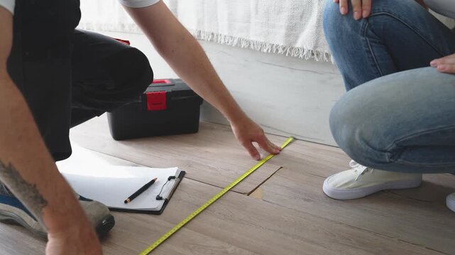 Professional handyman measuring damaged laminate flooring with tape measure while homeowner observes and contractor takes notes on clipboard, assessing repair needs and estimating project costs.
