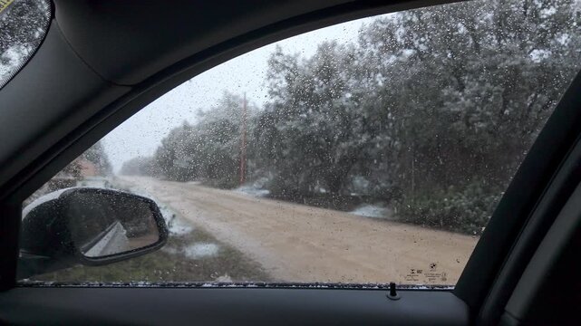 Slow motion snowfall seen through a wet car side window beside a snowy unpaved road and trees