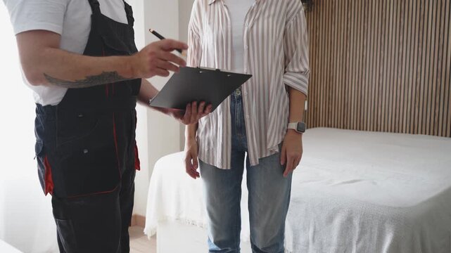 Young woman initially gesturing refusal, then discussing terms and finally signing a service agreement on a clipboard held by a professional handyman in overalls inside her bedroom