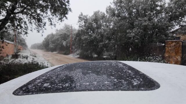 Slow motion snowfall over a snow covered car roof beside an unpaved road lined with trees
