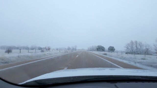 Vehicle point of view along a snowy rural road with frosted trees and falling snow in winter