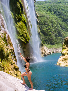 Woman stands barefoot, reaching toward waterfall