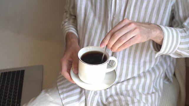 A man stirs coffee with a spoonful while taking a break while working remotely at home. The laptop is lying next to me on the sofa. Cozy home atmosphere, freelancing, relaxation during the working day