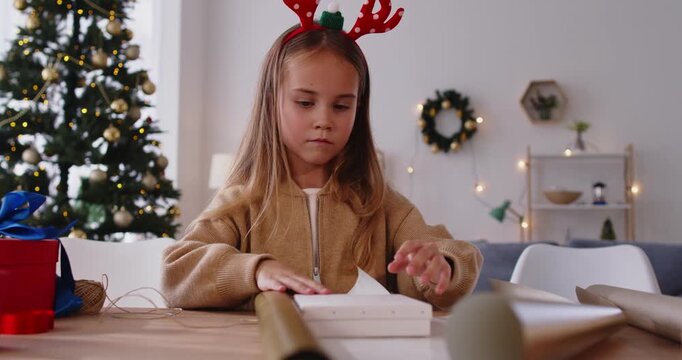 Christmas girl wrapping gift box in gold foil, sitting at home table beside Xmas tree. Child in reindeer headband using paper and thread on package, working in living room to prepare for holidays