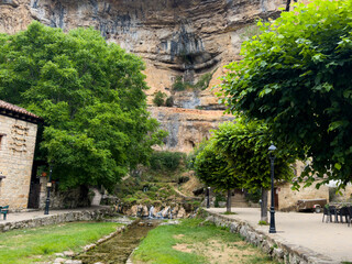 Fototapeta premium peaceful still scene of a small waterfall in a natural landscape with rocky cliff face and trees, captured from an eye-level perspective in an outdoor park or garden area with stone pathway.