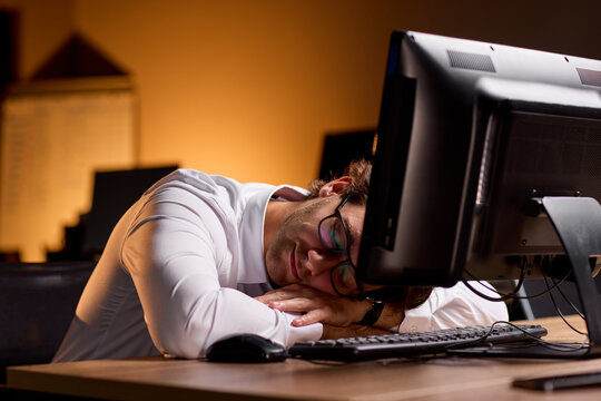 Stressed businessman rubbing eyes at desk in dim office after long workday, holding glasses with tired expression and warm blurred background during late evening