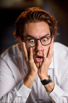 Young man in white shirt with glasses expressing shocked surprise holding face in hands under warm studio light, with man, young, surprised, shocked detail, showing expression, glass, hand, face