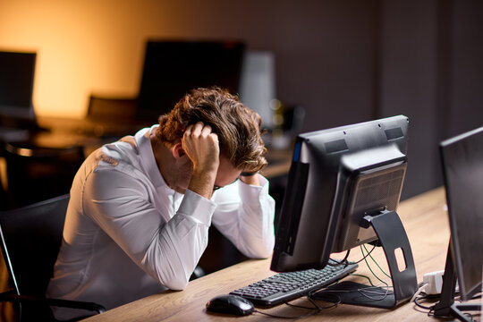 Stressed man holding head while sitting at office desk with computer late at night under warm lamp light, with man, stressed, fatigue, head detail, showing hand, office, desk, computer context