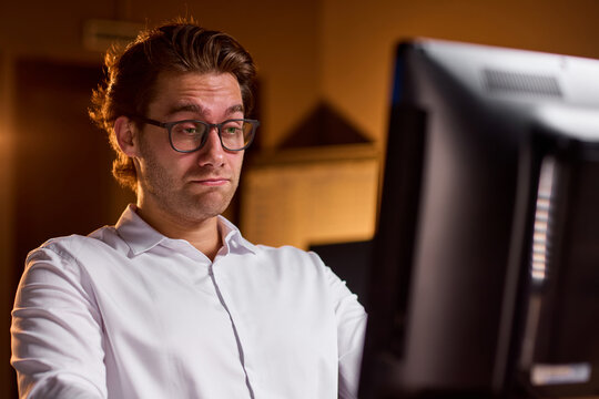 Stressed businessman rubbing eyes at desk in dim office after long workday, holding glasses with tired expression and warm blurred background during late evening