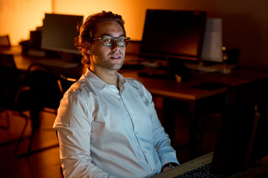 Stressed businessman rubbing eyes at desk in dim office after long workday, holding glasses with tired expression and warm blurred background during late evening