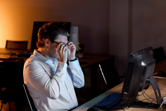 Tired office worker rubbing eyes at dim desk after long shift, computer screen glow on face, scattered chairs and keyboard nearby, exhausted mood, late night workplace stress