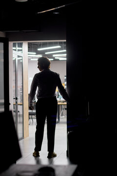 Silhouetted man opening glass door into brightly lit modern office interior at night, entering contemporary workplace with desks, chairs, overhead lighting and strong backlit contrast